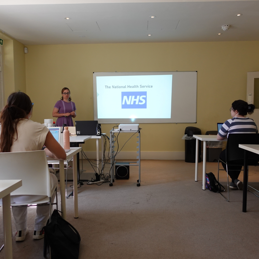 Claire Thompson, senior research fellow at the University of Hertfordshire stands at the front of a classroom next to a powerpoint that says NHS