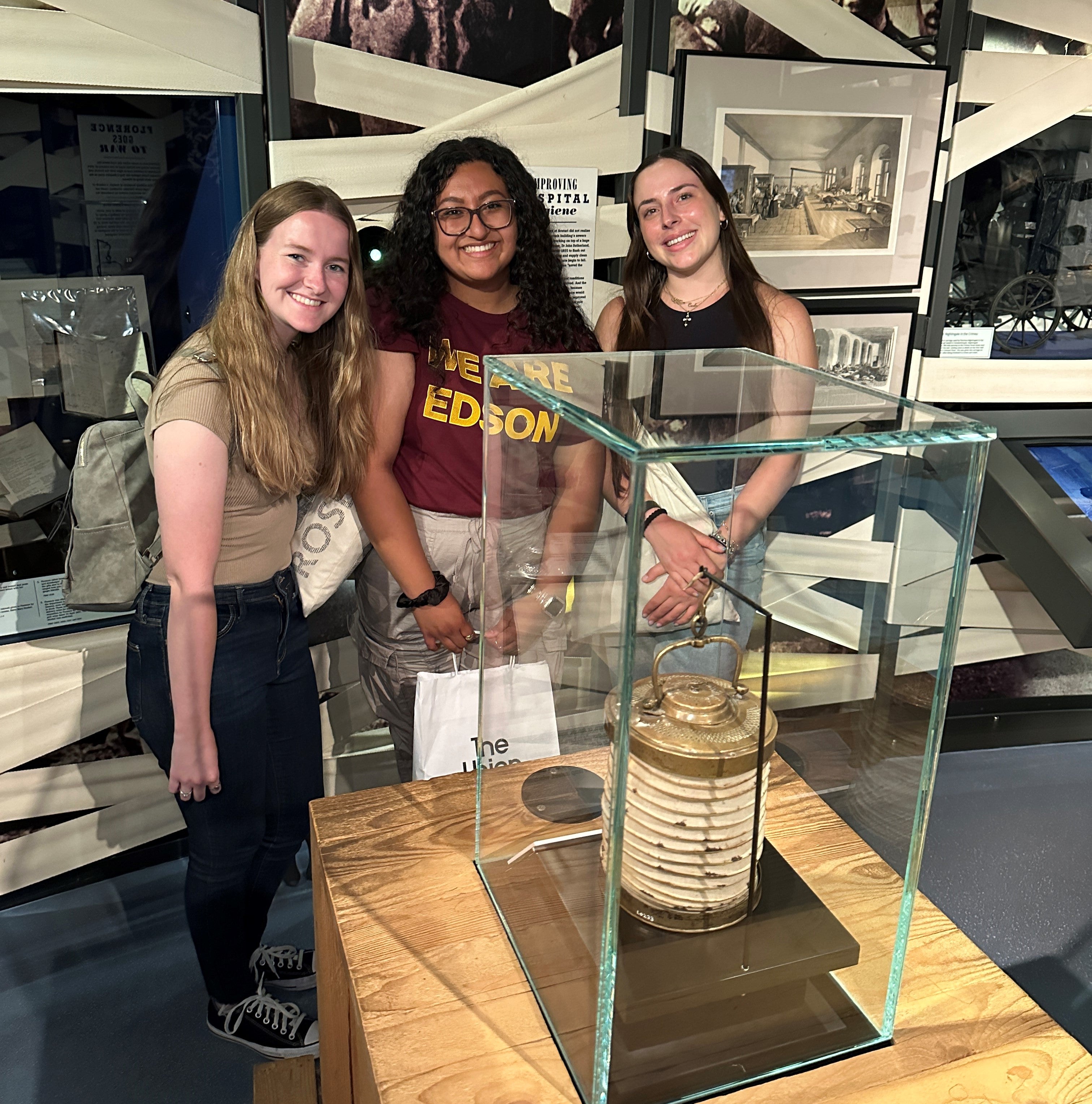 Three students pose in the Florence Nightingale Museum behind an oil lamp in a glass case