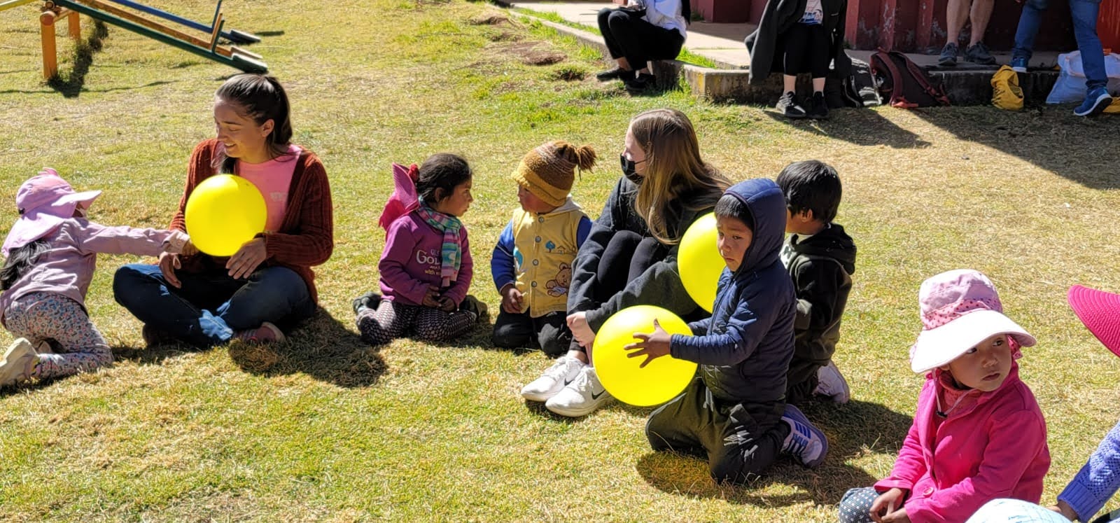 Students sit in a circle on the ground chatting with elementary age children in a community in Peru