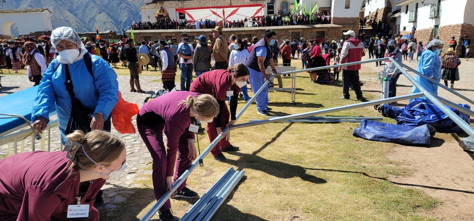 Students helping to set up a tent. They are holding metal poles to create the tent structure. In the background is a plaza full of Peruvian people