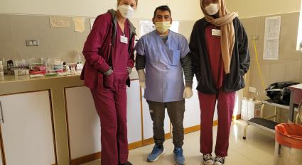 Nursing students in maroon scrubs stand on either side of a doctor wearing a blue scrub top inside of a medical clinic