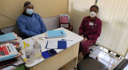 Shaniece Randolph sits next to a nurse in a clinic. She is wearing maroon scrubs and a surgical mask. The nurse is wearing a blue surgical gown and mask.