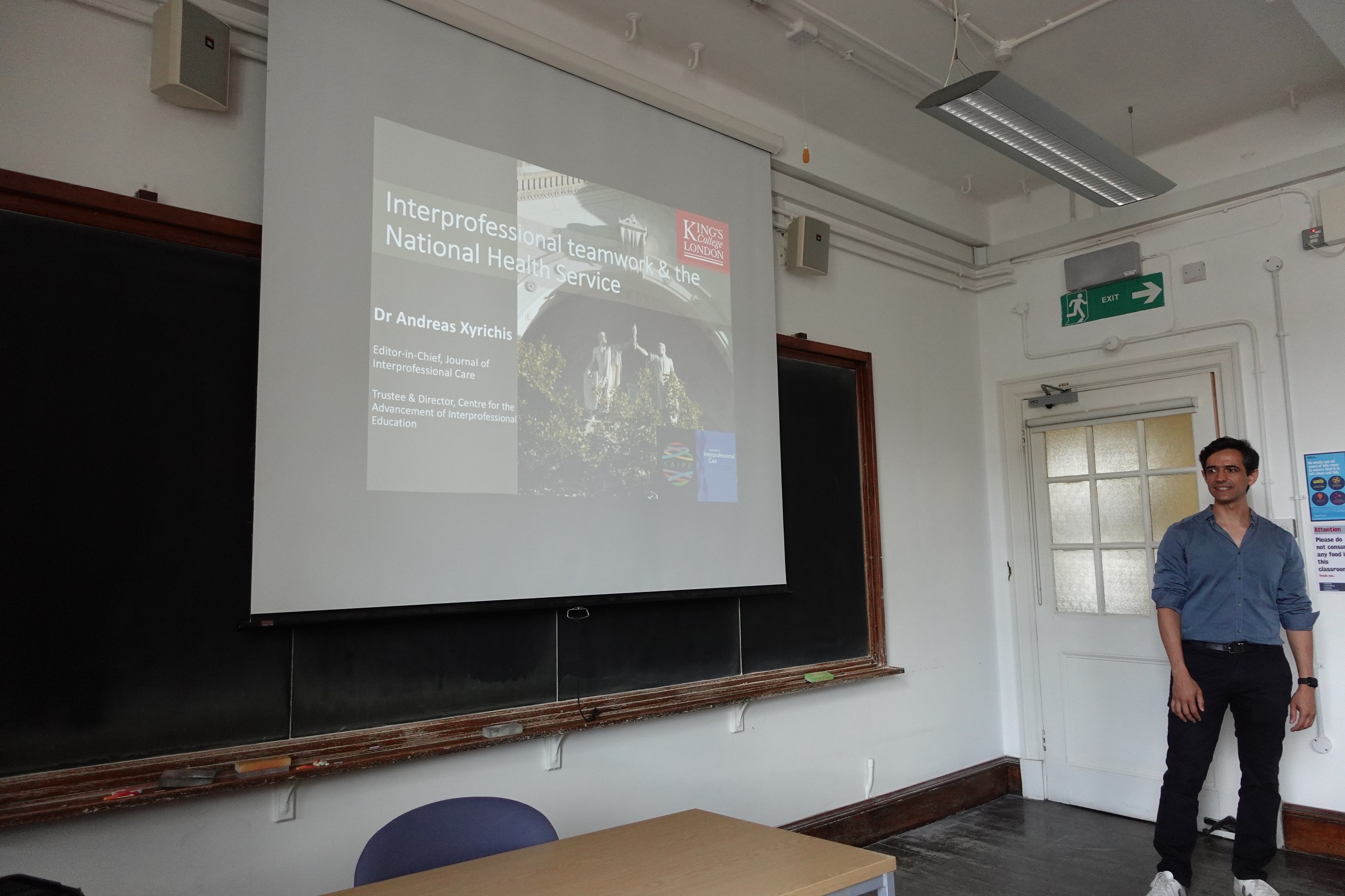 Andreas Xyrichis stands at the front of a classroom on the King's College London near a projector while presenting to students